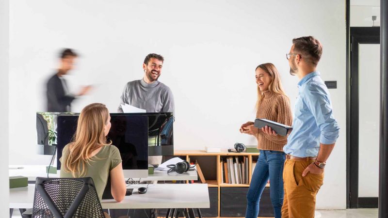 Team von jungen FachkrÃ¤ften im offenen BÃ¼ro, das sich lachend unterhÃ¤lt und Zusammenarbeit demonstriert.