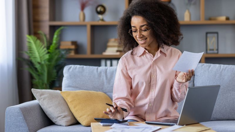 LÃ¤chelnde Person arbeitet eigenverantwortlich von zu Hause, Ã¼berprÃ¼ft Unterlagen und verwendet Laptop und Taschenrechner.