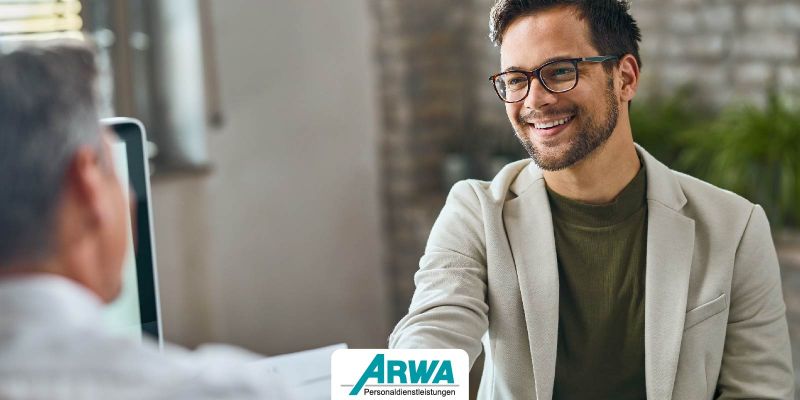 Ein junger Mann in einem beigen Blazer und mit Brille lächelt bei einem Vorstellungsgespräch mit einem Personalverantwortlichen in einem modernen Büro. Im Vordergrund das Logo von ARWA Personaldienstleistungen.