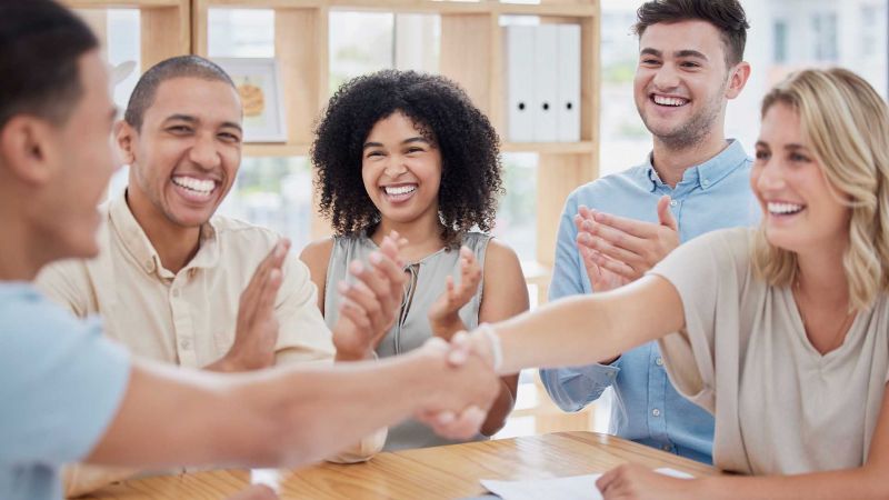 Eine Gruppe von fÃ¼nf lachenden Teammitgliedern applaudiert und schÃ¼ttelt sich die HÃ¤nde in einem modernen BÃ¼ro, was erfolgreiche Teamarbeit und Zusammenarbeit symbolisiert.