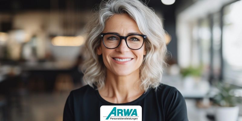 Professionell aussehende Frau mittleren Alters mit grauem Haar und Brille lächelt in einem modernen Büroraum, ARWA Personaldienstleistungen Logo im Vordergrund. Symbol für erfolgreiche Unternehmensführung.