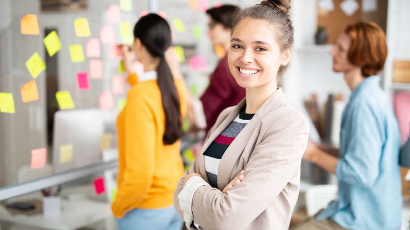 Eine junge Frau lÃ¤chelt in die Kamera, wÃ¤hrend im Hintergrund drei Teammitglieder an einem Whiteboard mit bunten Notizen arbeiten, was erfolgreiche Teamarbeit und Zusammenarbeit symbolisiert.