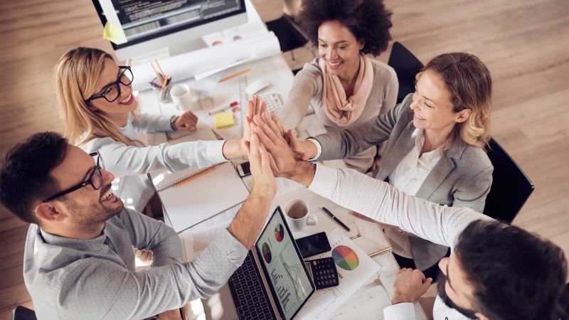 Ein Team von fÃ¼nf Mitarbeitern, die sich an einem Schreibtisch versammelt haben, gibt sich High-Fives und lÃ¤chelt. Auf dem Tisch liegen Dokumente und ein Laptop, die eine erfolgreiche Zusammenarbeit symbolisieren.