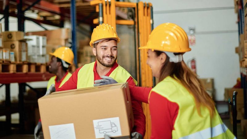 Zwei Lagerarbeiter mit gelben Schutzhelmen und Warnwesten arbeiten in einem Lager. Ein Mann Ã¼berreicht einer Frau eine Kartonbox, wÃ¤hrend ein weiterer Arbeiter im Hintergrund zu sehen ist. Sie lÃ¤cheln und scheinen eine gute Zusammenarbeit zu haben.