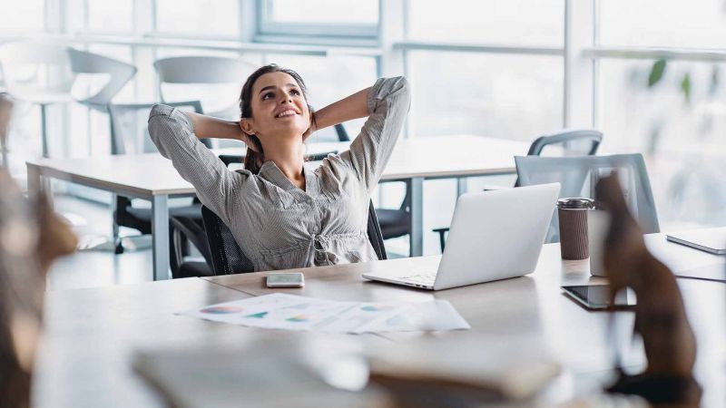 Frau lehnt sich entspannt im BÃ¼rostuhl zurÃ¼ck und lÃ¤chelt, symbolisiert StressbewÃ¤ltigung bei der Arbeit.