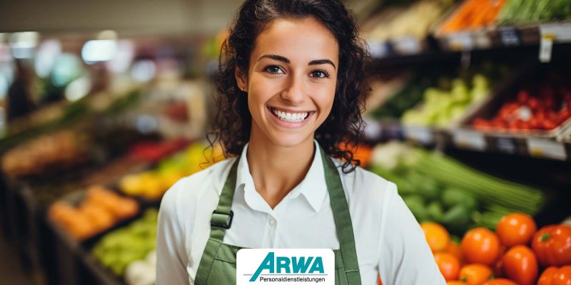 Lächelnde Frau in grüner Schürze arbeitet im Obst- und Gemüsebereich eines Supermarkts. ARWA Personaldienstleistungen Logo im Vordergrund.