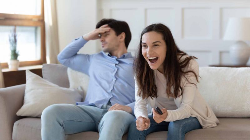 Eine Frau lacht laut auf, wÃ¤hrend ein Mann neben ihr auf dem Sofa sitzt und frustriert die Stirn hÃ¤lt, symbolisierend ambivalente Emotionen in einer Beziehung.