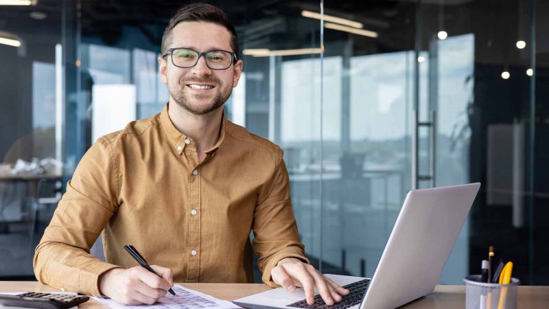 LÃ¤chelnder Mann mit Brille arbeitet am Laptop im BÃ¼ro, zeigt die StÃ¤rke der Introvertiertheit in einer ruhigen Arbeitsumgebung.