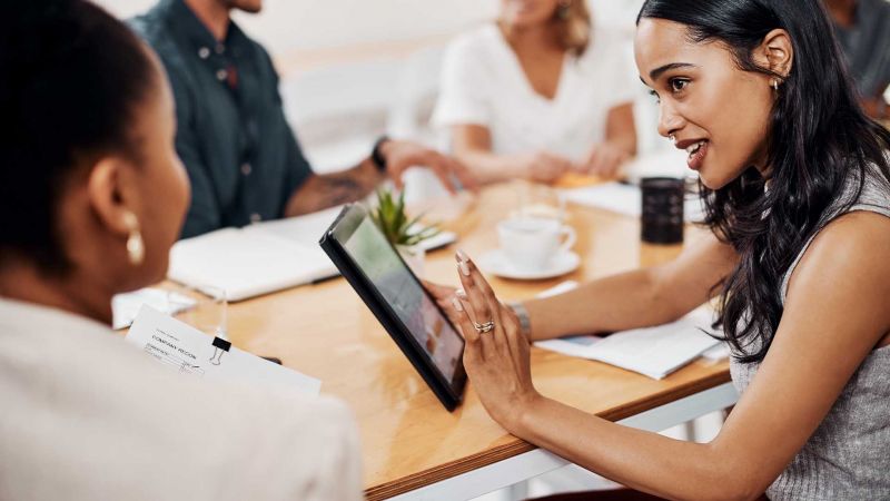 Zwei Frauen in einem geschÃ¤ftlichen Meeting, die eine zeigt etwas auf einem Tablet, wÃ¤hrend die andere zuhÃ¶rt, was Zusammenarbeit und Belastbarkeit im Beruf symbolisiert.