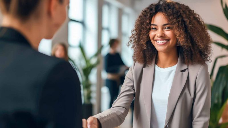 Junge Frau im Business-Outfit lÃ¤chelt und schÃ¼ttelt in einer BÃ¼roumgebung die Hand einer anderen Person, symbolisiert Vertrauensaufbau und Verantwortungsbewusstsein im Beruf.