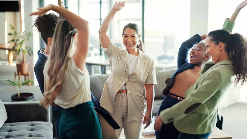 Gruppe von frÃ¶hlichen Frauen, die in einem BÃ¼ro tanzen und feiern, symbolisiert positive Energie und Teamgeist.