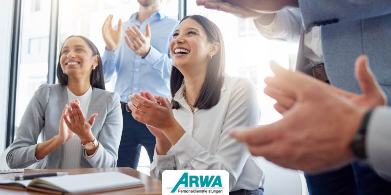 Gruppe von Menschen applaudiert in einem modernen Büro. Zwei Frauen im Vordergrund lächeln und klatschen, während sie motiviert an einem Meeting teilnehmen. ARWA Personaldienstleistungen-Logo im Vordergrund.