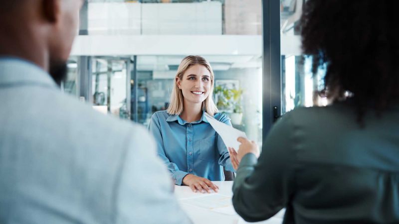 Bewerberin im VorstellungsgesprÃ¤ch, lÃ¤chelnd und mit Bewerbungsunterlagen, gegenÃ¼ber zwei Interviewern im modernen BÃ¼ro.