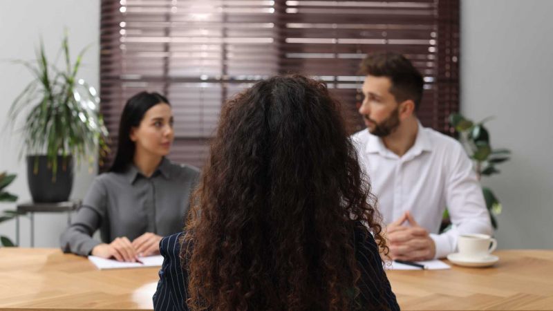 Frau sitzt vor zwei Interviewern in einem Besprechungsraum.