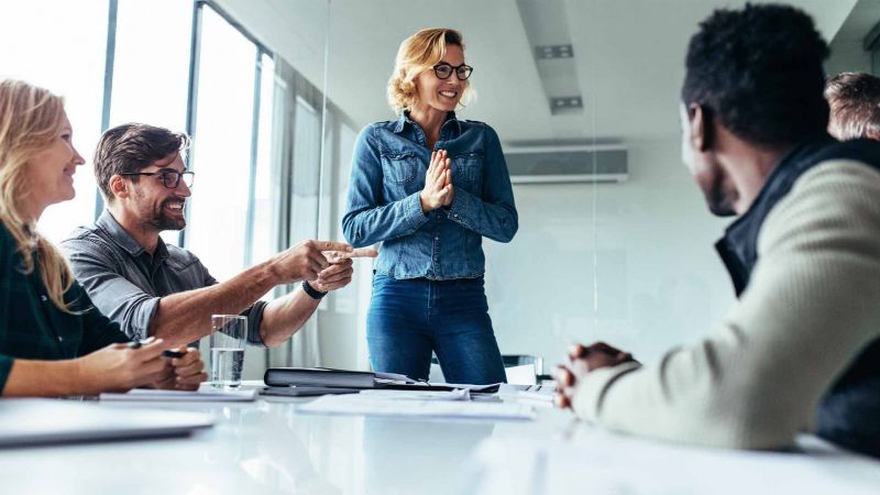 Eine Frau spricht in einem Teammeeting, wÃ¤hrend die anderen Teilnehmer durch LÃ¤cheln und Gestik ihre paraverbale Kommunikation unterstÃ¼tzen.