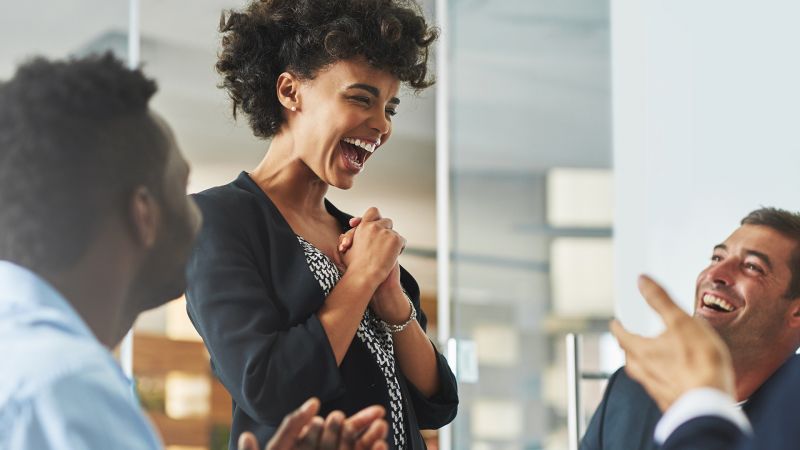 Begeisterte Frau im BÃ¼ro, die von ihren Kollegen Applaus erhÃ¤lt, symbolisierend hohe Mitarbeitermotivation und Anerkennung.