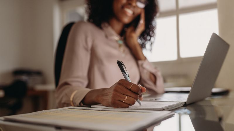 Frau an ihrem Schreibtisch, die lÃ¤chelnd telefoniert und Notizen macht, symbolisierend Mitarbeitermotivation und ProduktivitÃ¤t.