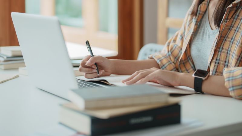 Junge Frau macht Notizen und arbeitet an einem Laptop, umgeben von BÃ¼chern, symbolisierend Wissbegierde und Lernbereitschaft.