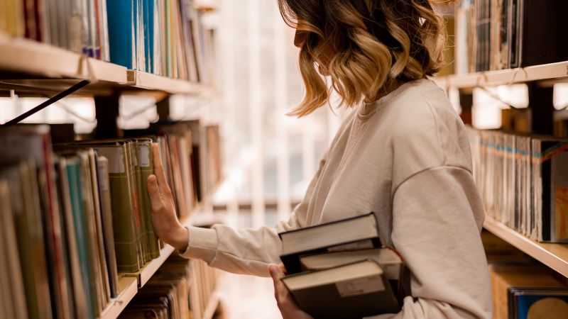 Junge Frau in einer Bibliothek, die nach BÃ¼chern greift, symbolisierend Wissbegierde und den Drang, neues Wissen zu entdecken.