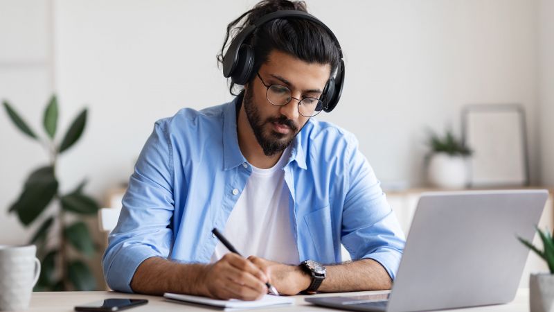 Junger Mann mit KopfhÃ¶rern, der konzentriert Notizen macht und dabei an einem Laptop arbeitet, symbolisierend Wissbegierde und Lernbereitschaft.