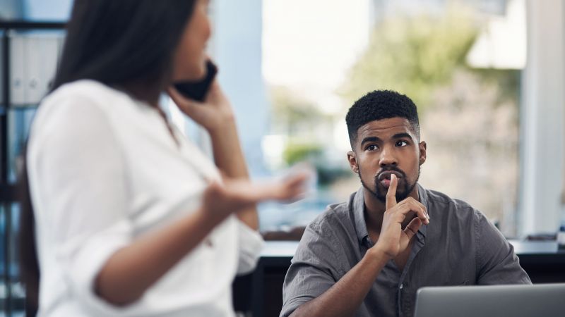 Ein Mann zeigt mit einer Geste zur Stille, wÃ¤hrend eine Frau gestikulierend telefoniert â€“ KÃ¶rpersprache im Arbeitsalltag.