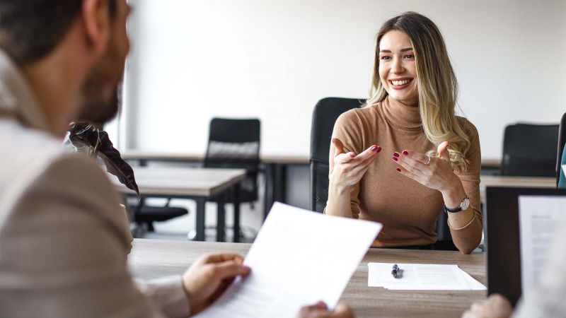 Eine junge Frau im VorstellungsgesprÃ¤ch beantwortet die Frage â€žWo sehen Sie sich in 5 Jahren?â€œ mit einem LÃ¤cheln und gestikuliert dabei engagiert.