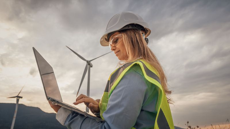 Ingenieurin mit Schutzhelm und Warnweste arbeitet mit einem Laptop vor einer Kulisse von Windkraftanlagen, symbolisiert nachhaltige Technologie und InnovationsfÃ¤higkeit.