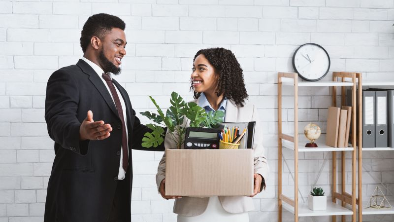 Zwei Personen in einem BÃ¼ro, eine Frau mit einer Kiste voller Arbeitsmaterialien und ein Mann, der sie freundlich begrÃ¼ÃŸt â€“ symbolisiert den Einstieg als Quereinsteiger.